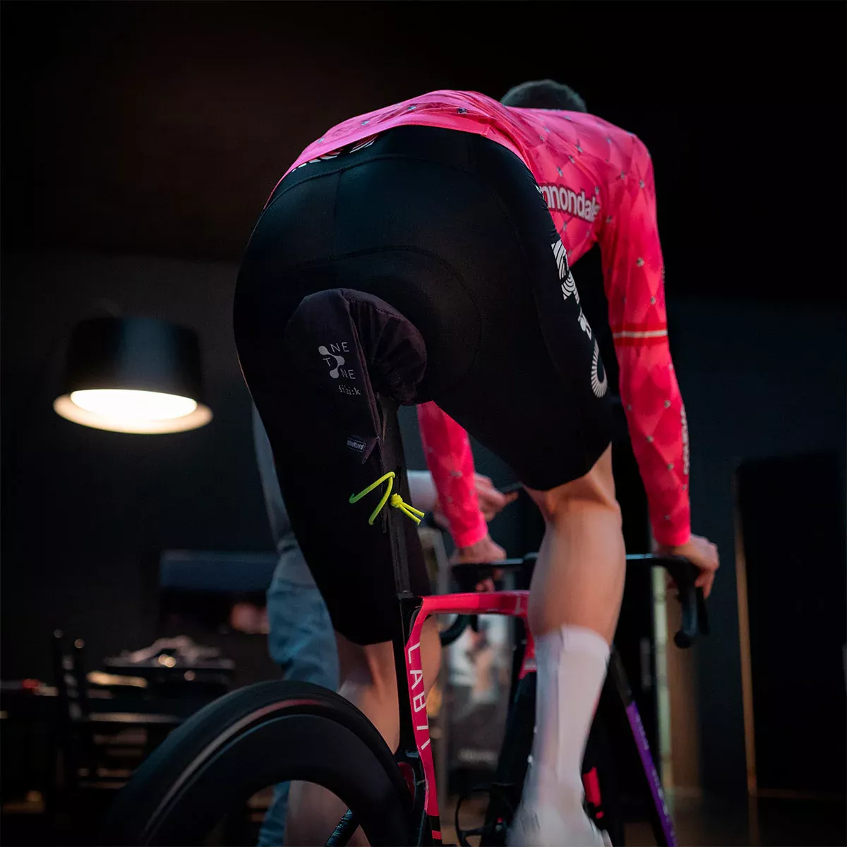 Ein Radfahrer in einem leuchtend rosafarbenen Trikot und schwarzen Shorts fährt in einem Innenraum auf einem stationären Fahrrad, von hinten gesehen. Der Raum ist schwach beleuchtet, im Hintergrund sind eine Lampe und einige Möbel zu sehen.