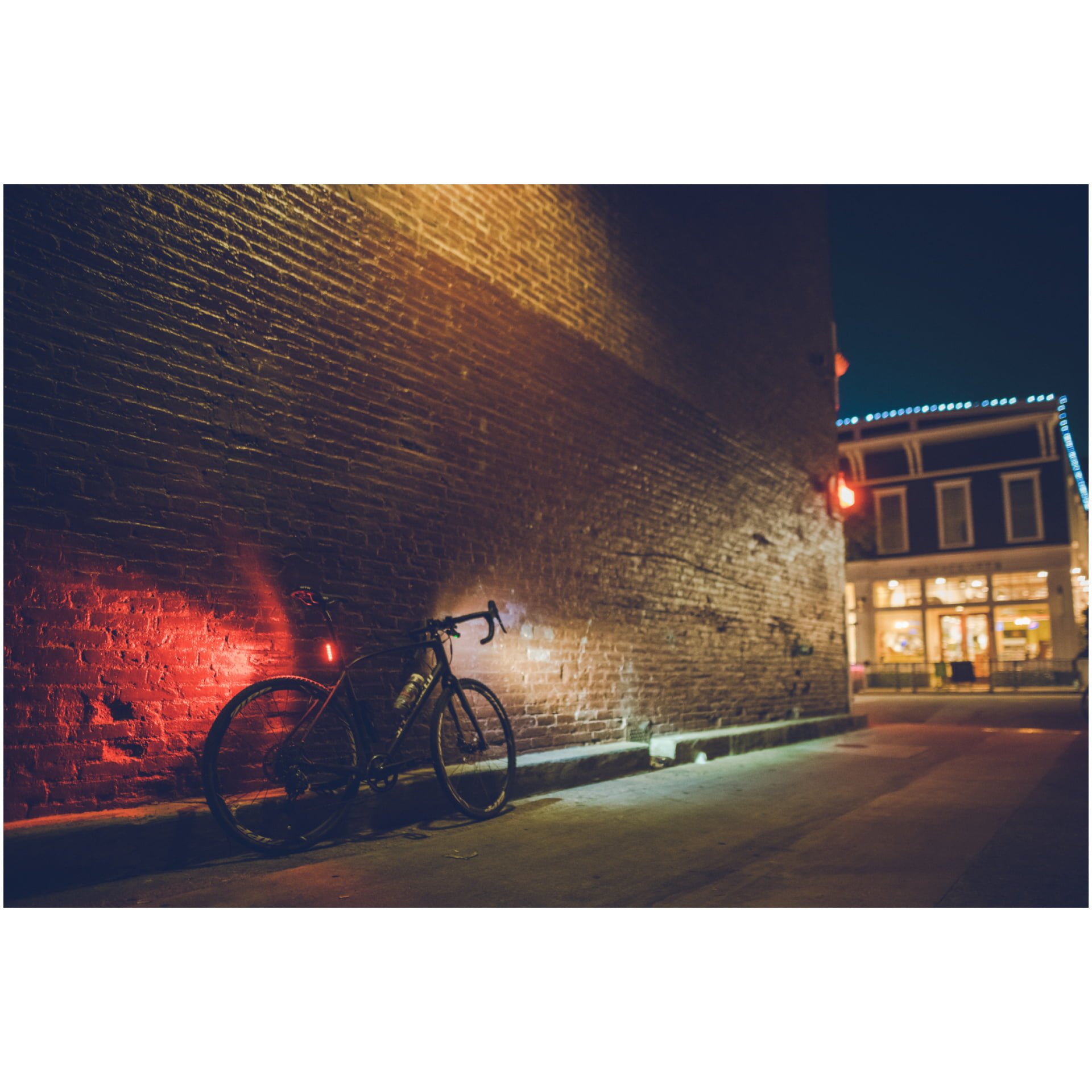 Ein Lucas-Fahrrad mit einem roten Rücklicht von Lezyne Strip Drive StVZO lehnt an einer Backsteinmauer in einer schwach beleuchteten Gasse. Die Gasse führt zu einem hell erleuchteten Gebäude im Hintergrund, dessen Dachlinie mit Lichterketten bestückt ist. Die gesamte Szenerie strahlt eine ruhige, nächtliche Atmosphäre aus.