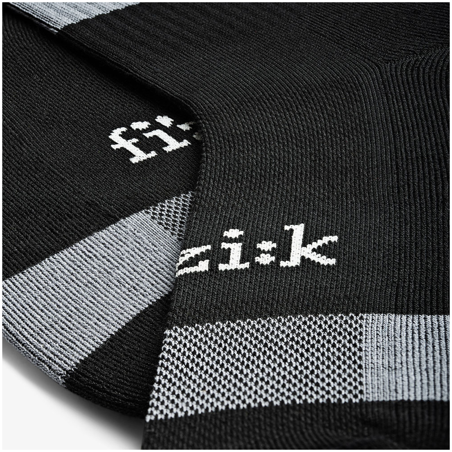 Nahaufnahme eines Paars FIZIK SOCKEN MTB. Die schwarzen und grauen Socken haben ein minimalistisches Design mit dem Markennamen „Fizik“, der in Weiß auf den schwarzen Stoff gestrickt ist. Die grauen Bereiche weisen ein strukturiertes Muster auf, das einen stilvollen Kontrast zum monochromen Thema bildet.