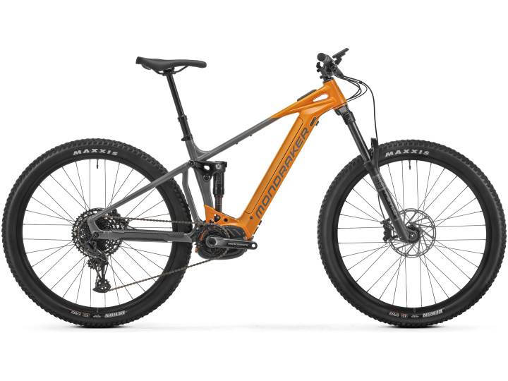 Das Mondraker Chaser ist ein Mountainbike mit einem orange-grauen Rahmen, der mit großen Maxxis-Reifen, einem Federungssystem und einem Sattel ausgestattet ist. Es verfügt außerdem über einen Elektromotor, der in der Nähe der Pedale angebracht ist, und trägt deutlich sichtbar die Aufschrift „Mondraker“ auf dem Rahmen.