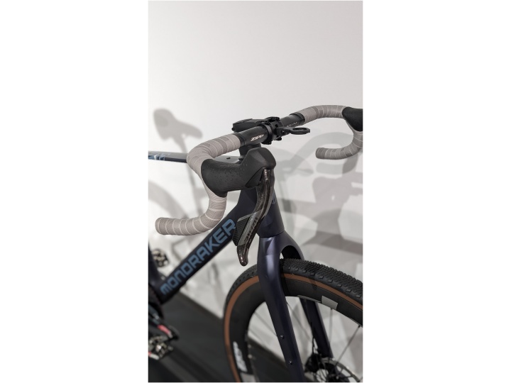 Eine Nahaufnahme des Mondraker Arid RR SL - L Lenkers zeigt umwickelte Griffe und Brems-Schalthebel, wobei ein Teil des Vorderrads und des Rahmens vor einem schlichten weißen Hintergrund zu sehen ist.