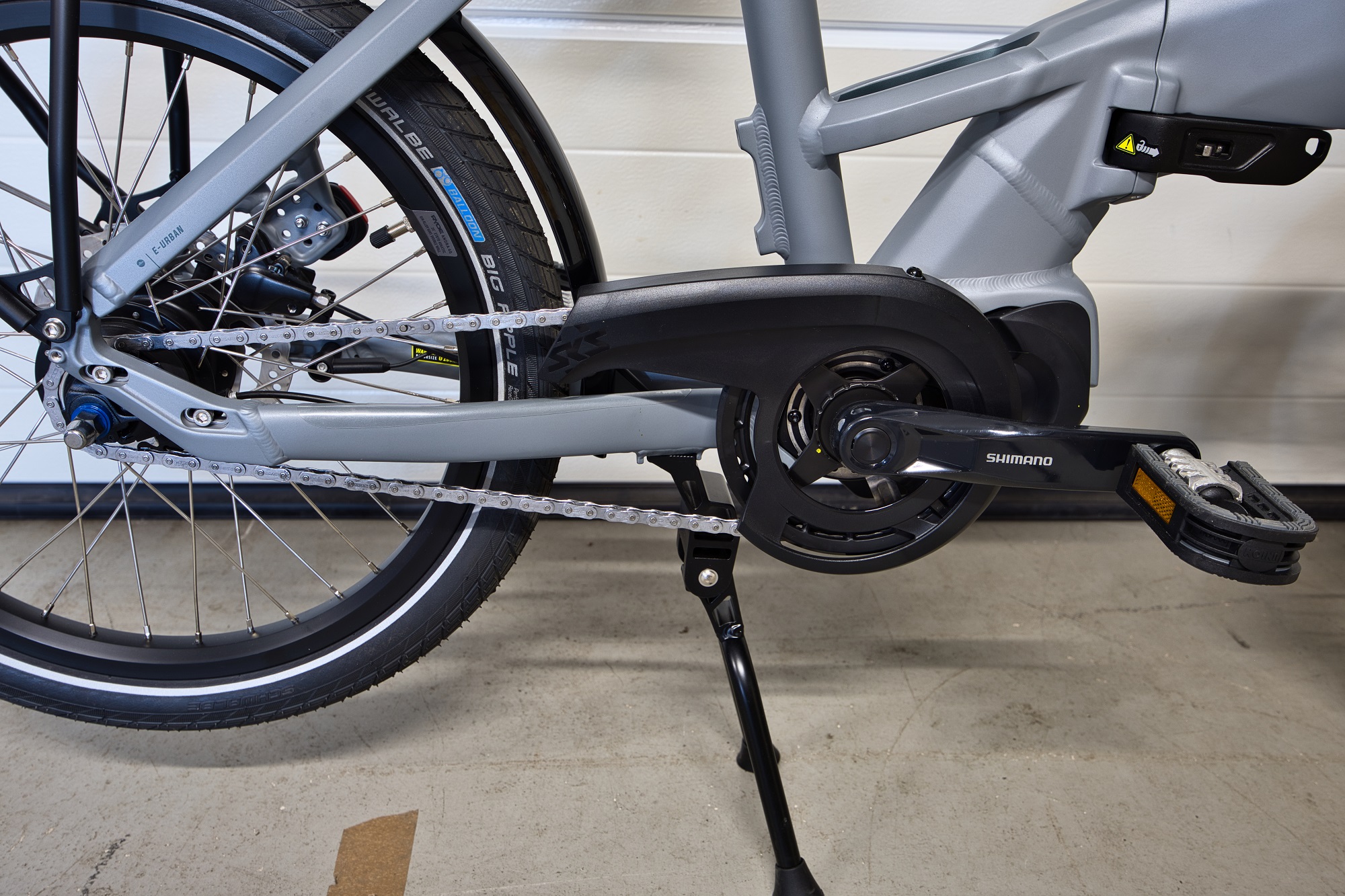 Nahaufnahme des Antriebsstrangs des Bergamont Paul-E EQ Edition mit einer Shimano-Kurbelgarnitur, einer Kette, einem Hinterrad und einem Ständer. Das E-Bike hat einen grauen Rahmen und schwarze Reifen und ist vor einem weißen Hintergrund abgebildet.